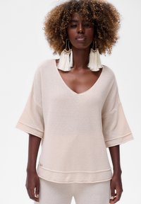 Vrouw met krullend haar, gekleed in een oversized beige gebreide top en bijpassende broek, geaccessorieerd met grote witte franjeringen tegen een witte achtergrond.