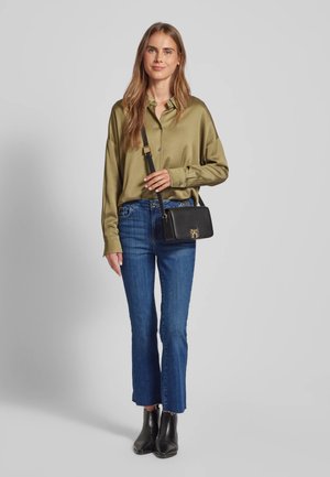 Camicia in raso verde oliva, jeans blu corti e stivaletti neri alla caviglia. Una piccola borsa nera con dettagli dorati è tenuta nella mano destra.