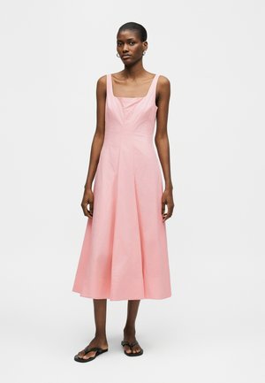 Kvinde iført en ærmeløs, lyserød midi-kjole med firkantet udskæring og sorte flade sandaler, poserende mod en ensfarvet lys baggrund.