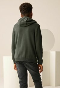 Dunkelgrüner, gestrickter Hoodie mit lockerem Sitz, der eine strukturierte Oberfläche und einen gerippten Saum aufweist, kombiniert mit schwarzen Jeans.