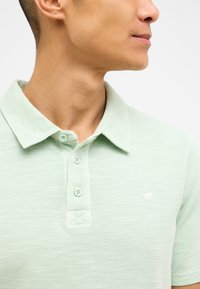 Jasnozielona koszulka polo z teksturowanej tkaniny, krótki rękaw, trzy guziki przy kołnierzy oraz małe haftowane logo po lewej stronie na piersi.