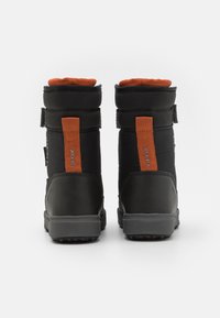 Botas impermeables negras con un cuello acolchado naranja, correa de velcro y una suela de goma; textura de tela y detalle de logo en la parte posterior.