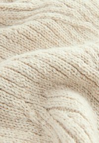 Beige strikket stof med et tekstureret zigzag-mønster. Materialet inkluderer subtile metalliske tråde, hvilket skaber et blødt og hyggeligt udseende.