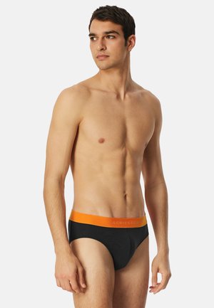 Nuori mies, jolla on lyhyet tummat hiukset ja joka pukeutuu mustiin bokseriin, jossa on oranssi vyötärönauha. Hän seisoo ja katsoo sivulle yksinkertaisen taustan edessä.