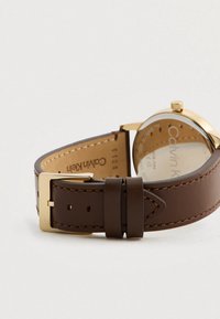 Bracelet de montre en cuir marron avec boucle en métal doré, présentant des coutures visibles et une finition lisse. Le fond du boîtier montre la marque et les détails.