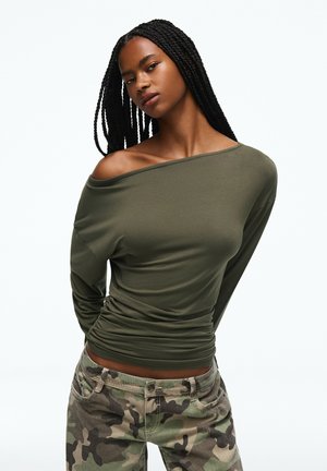 Femme aux longs cheveux tressés portant un haut vert olive épaules dénudées et un pantalon camouflage sur fond blanc uni.