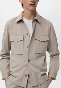 Beige button-up jacket with two chest pockets, a notch collar, and long sleeves. Made of smooth, lightweight fabric.
