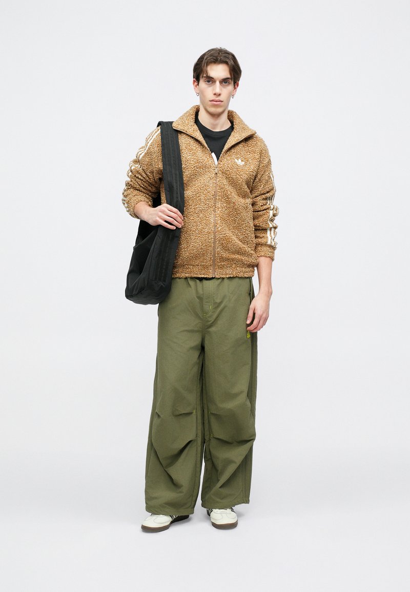 Brown fuzzy zip-up jacket with white stripes, paired with loose olive green pants. The model holds a black shoulder bag, wearing white sneakers.