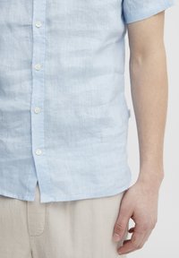 Camisa de lino de manga corta color azul claro con botón en la parte delantera, que presenta una tela texturizada y una etiqueta en la costura lateral.
