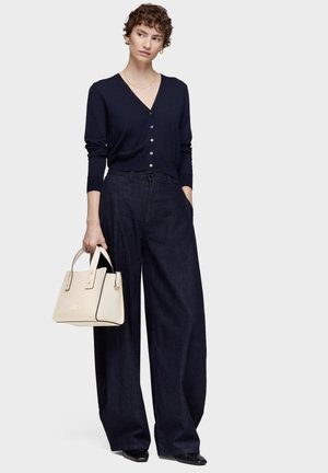 Donna con capelli corti e ricci che indossa un cardigan blu navy, jeans a gamba larga scuri, scarpe nere e tiene una borsa beige nella mano sinistra.