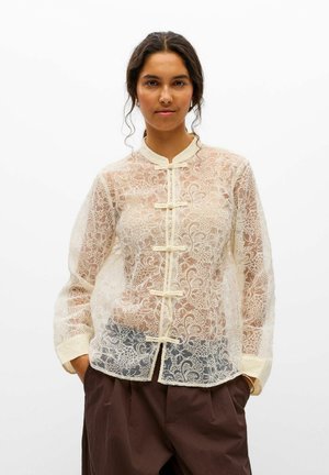 Un mannequin se tient devant un fond blanc, vêtu d’un chemisier en dentelle florale ivoire transparent, à fermetures à boucles, et d’un pantalon marron foncé.