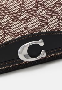 Brun och krämvävt tyg med cirkulära mönster, svarta läderdetaljer, silverfärgad logohardware och kontrasterande sömnadsdetaljer.