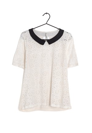 Blouse blanche en dentelle à manches courtes avec des motifs floraux et un col Peter Pan noir contrastant, suspendue à un cintre noir.