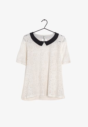 Blouse blanche en dentelle à manches courtes avec des motifs floraux et un col Peter Pan noir contrastant, suspendue à un cintre noir.