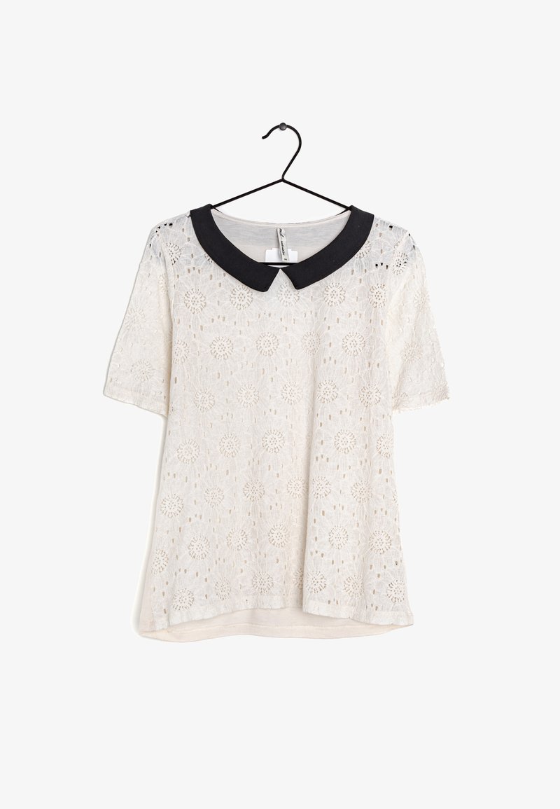 Blouse blanche en dentelle à manches courtes avec des motifs floraux et un col Peter Pan noir contrastant, suspendue à un cintre noir.