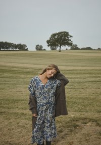 Blaues Kleid mit Blumenmuster, puffärmel und V-Ausschnitt, unter einem braunen Strick-Cardigan in einer grasbewachsenen Landschaft.