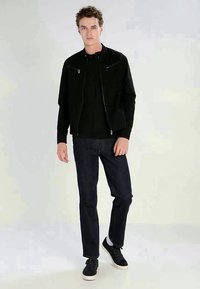 Melns bomber jakas ar rāvējslēdzēju priekšpusē, kopā ar melnu T-kreklu un tumšiem džinsiem. Modelis stāv, uzvilcis melnu kedu. Neitrāls fons.
