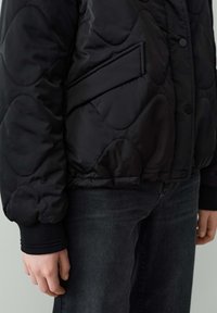 Veste matelassée noire avec un design arrondi, des boutons-pression et des poignets élastiques. Comprend une poche latérale et une texture lisse.
