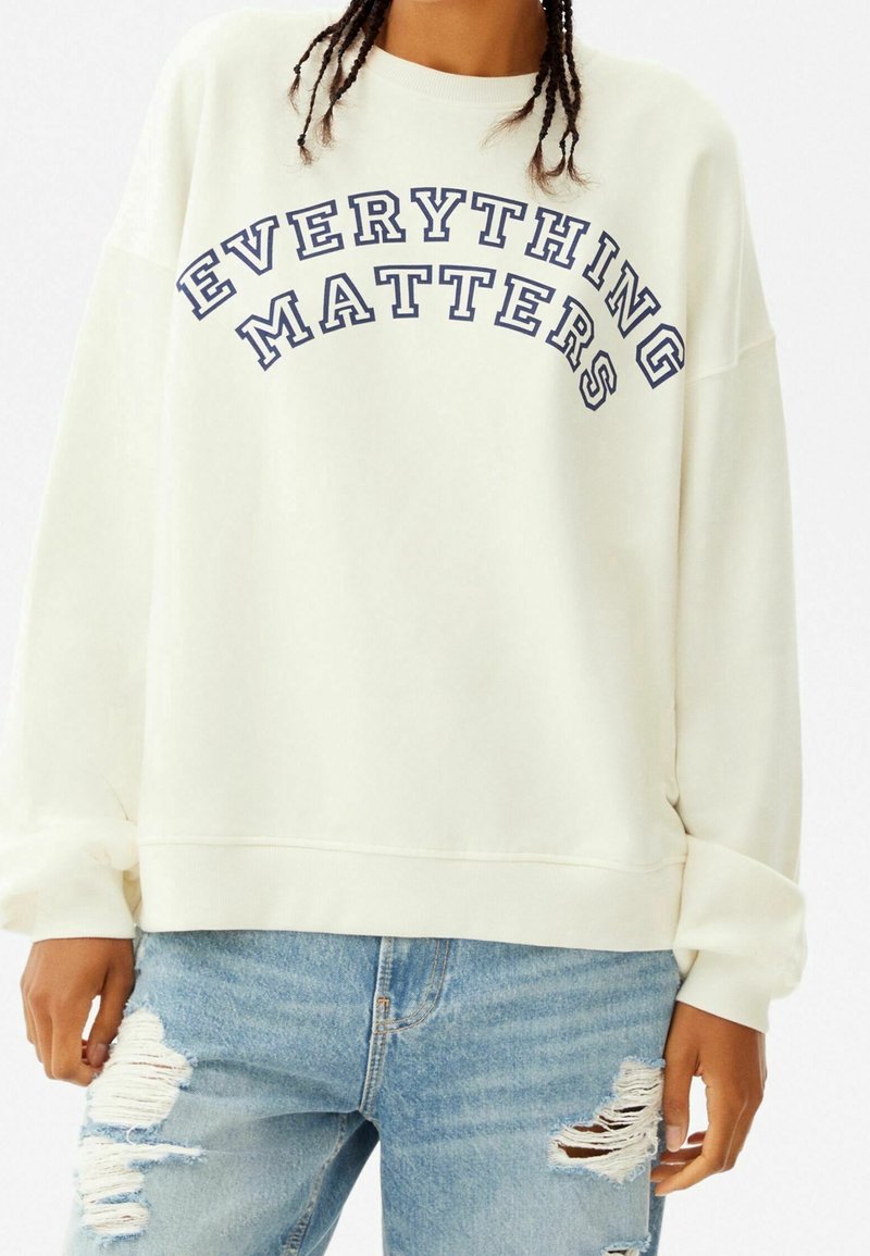 Krämfärgad sweatshirt med "EVERYTHING MATTERS" tryckt i mörkblå bågtext, med löst sittande ärmar och en texturerad yta.