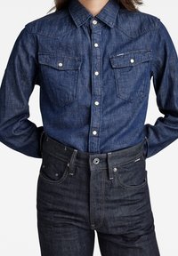 Persona que lleva una camisa de mezclilla de botones de manga larga de color azul oscuro metida dentro de unos jeans de mezclilla azul oscuro, con las manos detrás de la espalda.