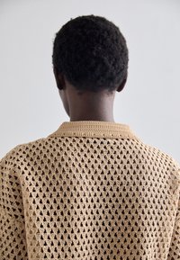 Strikket cardigan i beige med et hulmønster i trekantform, der har en rund krave og en løs pasform, som fremhæver en åndbar struktur.