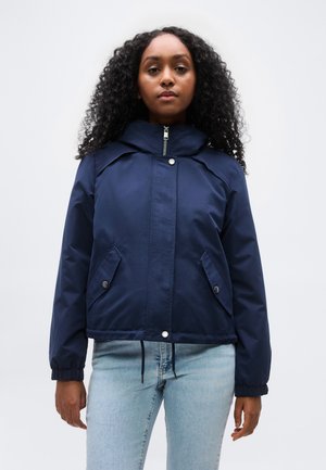 Femme aux cheveux bouclés portant une veste zippée bleu marine avec des poches à boutons-pression et un jean bleu clair, debout devant un fond uni.
