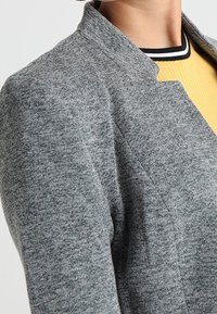 Šedá pletená bunda s texturovanou látkou, s límcem a čistými liniemi. Dole se rýsuje žlutý žebrovaný top s černými akcenty.