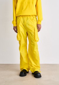 Pantaloni cargo gialli realizzati in tessuto leggero, dotati di tasche laterali, una vita elasticizzata e una vestibilità comoda, abbinati a scarpe nere.
