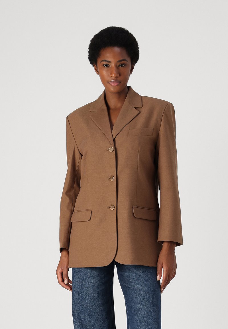 Brun blazer med en strukturert design, med noterte revers, fem knapper, to frontlommer og lange ermer, kombinert med blå jeans.