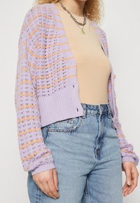 Cardigan en maille lilas avec motif à maille ouverte et accents orange. Dispose d'une coupe courte, de poignets côtelés et d'une fermeture à boutons. Porté sur un haut beige.