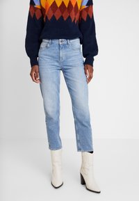 Jeans in denim azzurro chiaro con taglio dritto, indossati con un maglione a motivo multicolore e stivali bianchi con tacco alto.