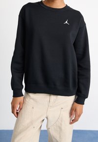 Sudadera negra de tejido suave, con cuello redondo acanalado y puños, y con un logo de Jordan en blanco en el pecho. Combinada con pantalones claros de color beige.