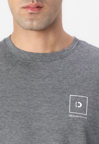 Gråstripete t-skjorte med rund hals, med en liten hvit logo i firkantet design på venstre side. Myk stofftekstur.