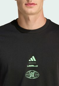 Camiseta de algodón negra con cuello redondo, que presenta un logo reflectante verde de Adidas y el texto "LesMills LIVE '25" impreso en la parte frontal.