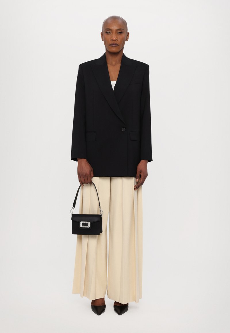 Blazer nero oversize, pantaloni plissettati color crema, décolleté nere a punta, piccola borsa nera con dettagli in cristallo. Tessuto liscio e silhouette strutturata.