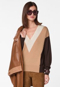 Beige und braun farbblockiertes Sweatshirt mit V-Ausschnitt. Begleitet von einer braunen Lederjacke mit breiten Revers und Knopfakzenten.