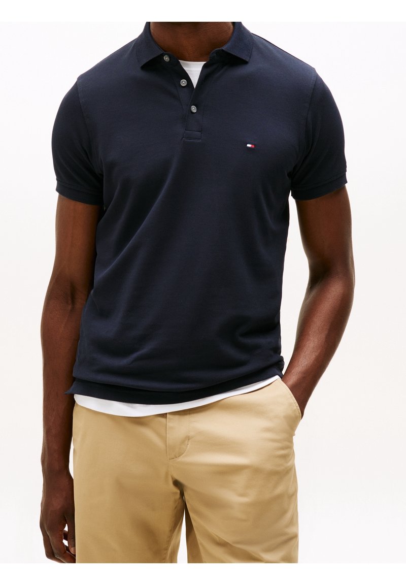 Mörkblå polo tröja i bomull med korta ärmar, krage med två knappar, lätt textur och en liten logotyp på bröstet. Kombinerad med beiga byxor.