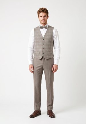 Man wearing beige plaid vest, white dress shirt, matching beige trousers, gray bow tie, and brown dress shoes standing against plain background.