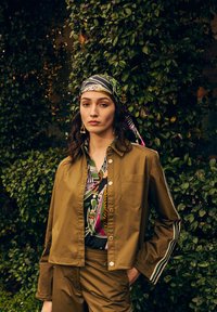 Olive green button-up jacket with white stripes, layered over a colorful patterned shirt and matching pants, paired with a patterned headscarf.