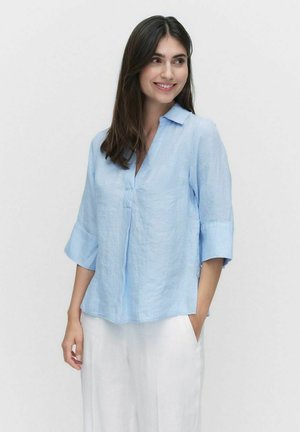Chemise en lin bleu clair avec un col en V, des manches trois-quarts et une coupe décontractée. Elle possède une poche sur le côté gauche et une texture douce.