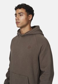 Un giovane con capelli corti e ricci, che indossa una felpa marrone con un piccolo logo sul petto, guarda verso sinistra su uno sfondo neutro.