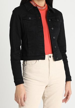 Frau trägt eine schwarze Jeansjacke mit Knöpfen über einem roten Hemd mit weißem Text am Kragen und cremefarbene, hochtaillierte Cordhosen.