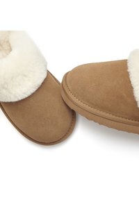 Tan suede slippers with a soft, cream-colored shearling cuff. Rounded toe, flat sole, and stitched edges visible.
