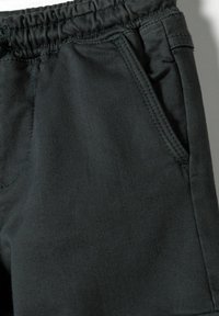Shorts en coton noir avec une taille élastique, une poche latérale et une texture lisse. Coutures visibles le long des bords et de la couture de la poche.