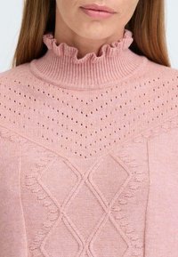 Pull en tricot rose avec un col haut ondulé à côtes. Le motif présente des textures en forme de diamant et de petites perforations partout.