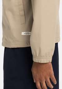 Beige Utility-Jacke mit einem gerippten Bündchen und einem Etikett am Saum. Der Stoff sieht glatt und leicht aus und wird mit dunklen Hosen kombiniert.