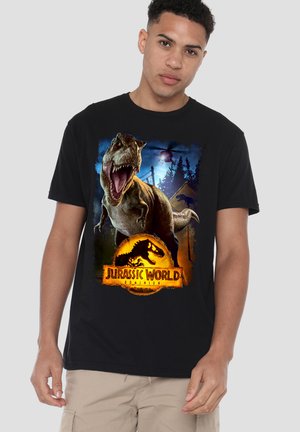 Junger Mann trägt ein schwarzes Jurassic World Dominion T-Shirt mit einem brüllenden T-Rex, Wald, Hubschrauber und Warnschild.