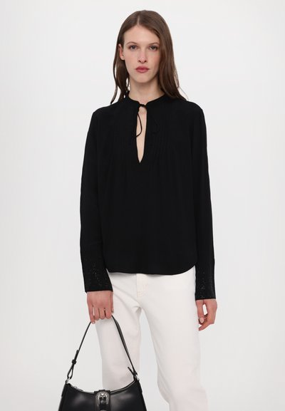 Black long-sleeve blouse with a keyhole neckline, pleated detailing, and textured sparkly cuffs, paired with white pants and a black handbag.