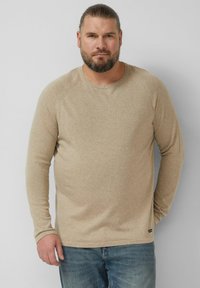 Beżowa koszulka z długim rękawem i okrągłym dekoltem, wykonana z miękkiego materiału, o luźnym kroju i subtelnej fakturze. Połączona z niebieskimi jeansami.