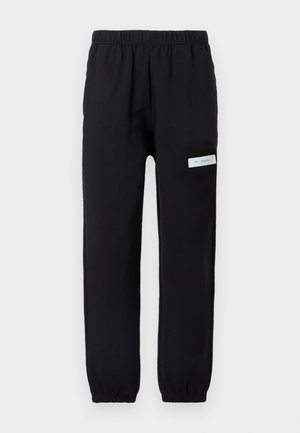 Pantaloni de trening negri cu talie elastică, picioare conice și o etichetă albă pe partea stângă. Textură netedă, design casual.